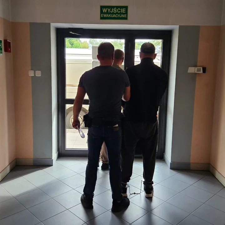 Trzej mężczyźni, w tym jeden w kajdankach, stoją przy drzwiach wyjścia ewakuacyjnego budynku.