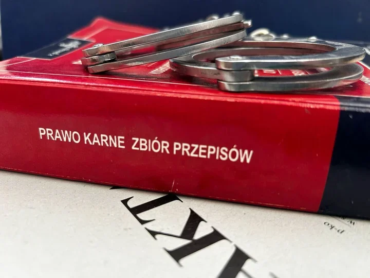 Kajdanki leżą na czerwonym tomie zbioru przepisów prawa karnego, a pod książką widać fragment dokumentu z dużymi literami.