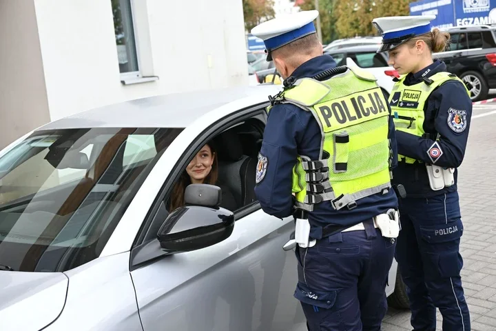 Dwóch policjantów drogówki rozmawia z kierującą samochodem osobowym na parkingu przed budynkiem.