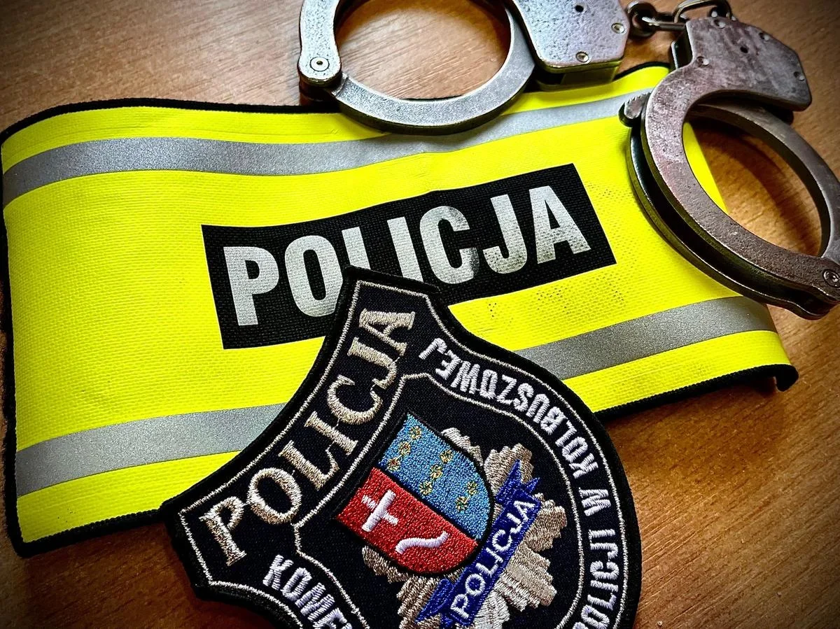 Żółta opaska z napisem POLICJA, policyjna naszywka i kajdanki leżą na drewnianej powierzchni.