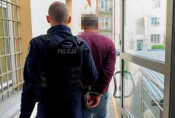 Funkcjonariusz policji prowadzi zakutego w kajdanki mężczyznę korytarzem budynku.