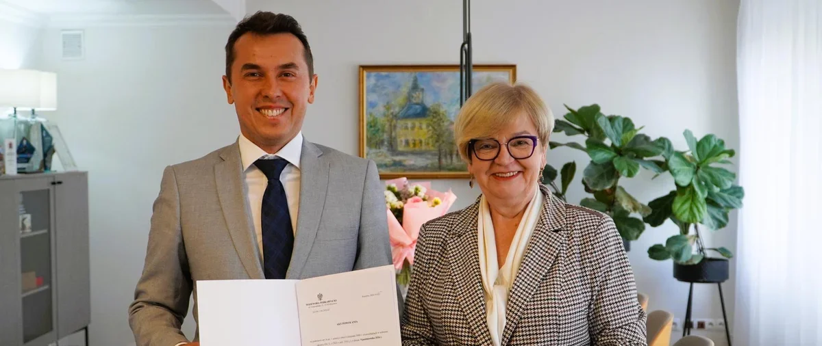 Mężczyzna w szarym garniturze trzyma otwarty dokument, stojąc obok kobiety w pepitce, w jasnym pomieszczeniu biurowym z obrazem i rośliną w tle.