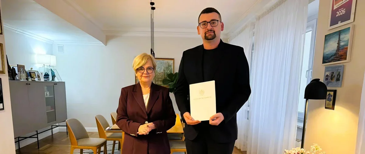 Mężczyzna w czarnym garniturze trzyma dokument z godłem, stojąc obok kobiety w bordowym garniturze w elegancko urządzonym wnętrzu mieszkalnym.