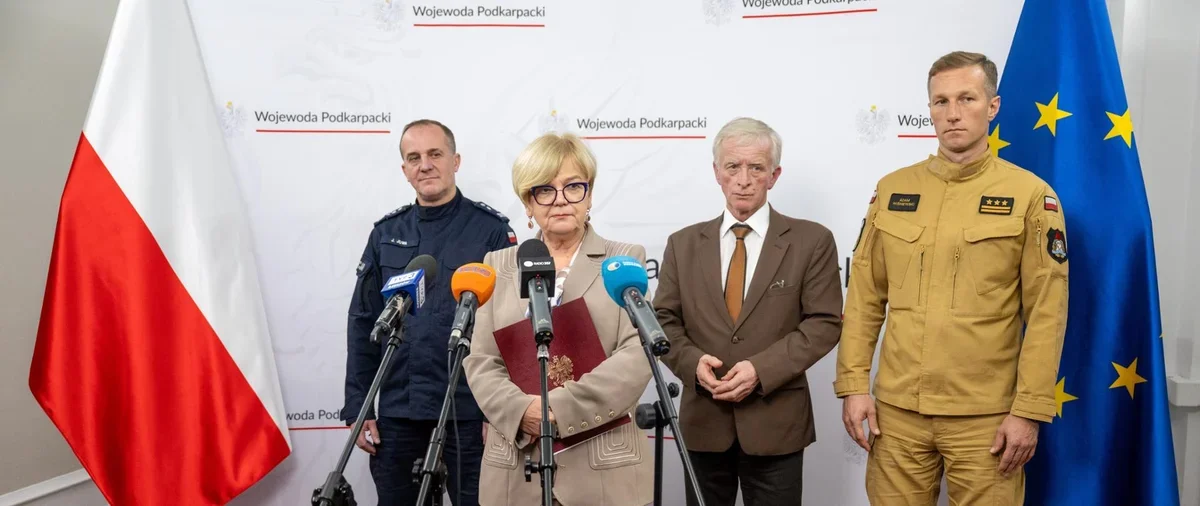 Cztery osoby, w tym funkcjonariusze służb mundurowych i osoba cywilna, stoją podczas konferencji prasowej przed flagami Polski i UE oraz banerem Wojewody Podkar