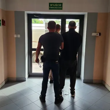 Trzej mężczyźni, w tym jeden w kajdankach, stoją przy drzwiach wyjścia ewakuacyjnego budynku.