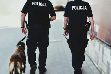 Dwóch policjantów w mundurach z napisem POLICJA idzie brukowaną alejką, jeden prowadzi psa służbowego na smyczy.