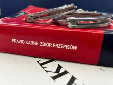 Kajdanki leżą na czerwonym tomie zbioru przepisów prawa karnego, a pod książką widać fragment dokumentu z dużymi literami.