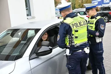 Dwóch policjantów drogówki rozmawia z kierującą samochodem osobowym na parkingu przed budynkiem.
