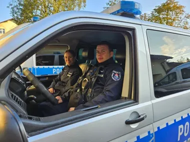Dwójka policjantów w mundurach siedzi w kabinie oznakowanego radiowozu policyjnego.