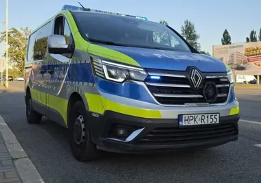 Policyjny oznakowany radiowóz Renault Trafic z tablicą HPK R155 stoi na poboczu drogi w słoneczny dzień.