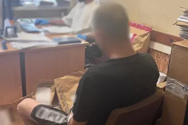 Mężczyzna w czarnej koszulce siedzi w pomieszczeniu biurowym przy biurku, na którym leżą dokumenty i sprzęt, a w tle widoczna jest druga osoba.