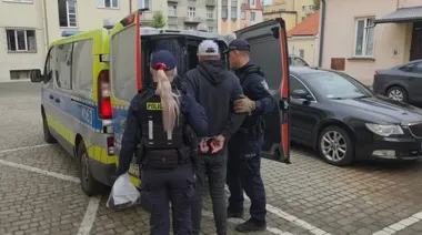 Funkcjonariusze policji prowadzą zatrzymanego mężczyznę w kajdankach do tyłu policyjnego samochodu typu furgonetka na brukowanym parkingu w zabudowie miejskiej.