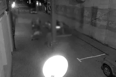 Nocne zdjęcie z monitoringu przedstawiające rozmyte postacie ludzi na brukowanej uliczce między budynkami, z widocznym samochodem i jasnym światłem w pierwszym