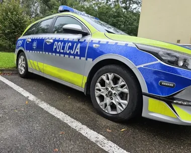 Policyjny radiowóz marki Kia z napisem „Policja” stoi zaparkowany na asfaltowej nawierzchni przy białej linii, w tle widać zieleń i fragment budynku.