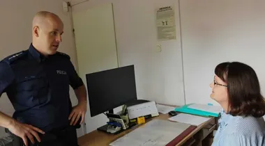 Policjant w granatowym mundurze rozmawia z kobietą siedzącą przy biurku w pomieszczeniu biurowym.