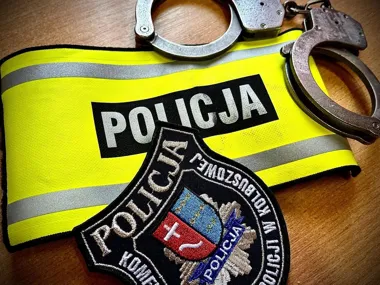 Żółta opaska z napisem POLICJA, policyjna naszywka i kajdanki leżą na drewnianej powierzchni.