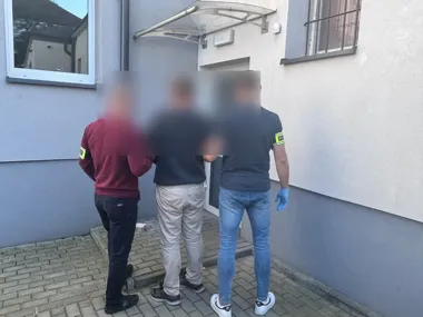 Dwaj mężczyźni z opaskami policyjnymi prowadzą zatrzymanego mężczyznę przed wejściem do budynku.