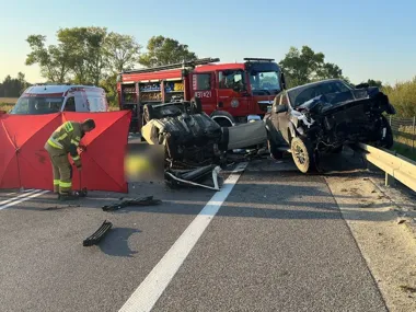 Strażak ustawia parawan przy wrakach dwóch rozbitych samochodów na drodze, w tle wóz strażacki i karetka pogotowia.