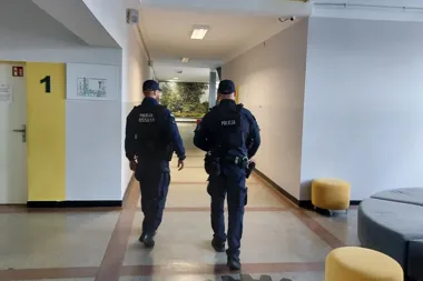 Dwóch policjantów w mundurach przechodzi korytarzem budynku publicznego, obok żółtej ściany z numerem 1.