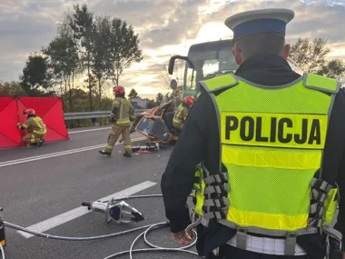 Policjant obserwuje akcję strażaków przy rozbitym pojeździe na drodze o zachodzie słońca.