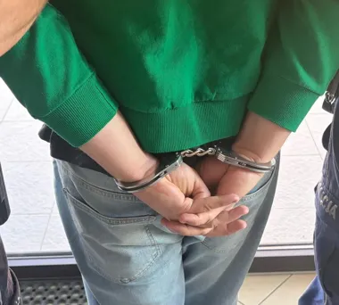 Mężczyzna w zielonej bluzie i jeansach stoi z rękami skręconymi za plecami w kajdankach.