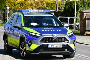Policyjny radiowóz marki Toyota z napisem „POLICJA” i numerem rejestracyjnym HPK A060 jedzie po asfaltowej drodze w słoneczny dzień.
