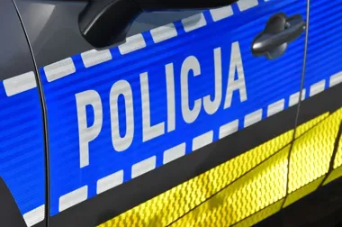Fragment boku policyjnego radiowozu z napisem POLICJA, niebiesko-białą szachownicą i żółtym pasem odblaskowym.
