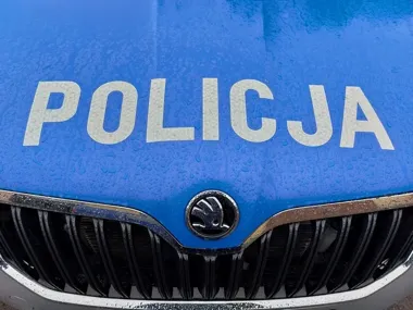 Fragment niebieskiego policyjnego radiowozu marki Škoda z napisem POLICJA na masce, pokrytej kroplami wody.