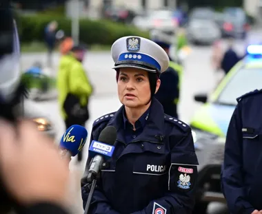 Policjantka w mundurze udziela wywiadu dziennikarzom na ulicy, w tle radiowóz i inni funkcjonariusze.