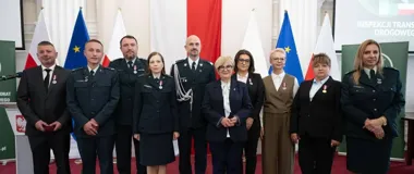 Grupa osób, w tym funkcjonariusze Inspekcji Transportu Drogowego i urzędnicy, pozuje do pamiątkowego zdjęcia podczas uroczystości w sali z flagami polskimi i un