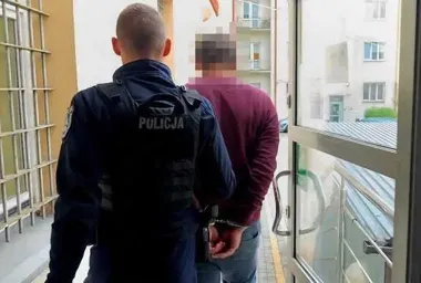 Funkcjonariusz policji prowadzi zakutego w kajdanki mężczyznę korytarzem budynku.
