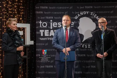 Trzy osoby stoją na scenie przed ścianką z napisami Teatru Rzeszowskiego podczas oficjalnej uroczystości.