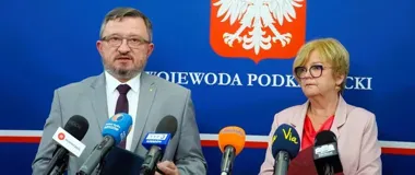Mężczyzna w garniturze przemawia do mikrofonów podczas konferencji prasowej, obok niego stoi kobieta, w tle herb i napis „Wojewoda Podkarpacki”.