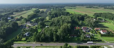Widok z lotu ptaka na zabudowę jednorodzinną w otoczeniu pól i lasów, przeciętą drogą krajową z pojazdami.