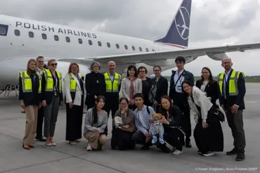 Grupa osób, w tym pracownicy lotniska w kamizelkach odblaskowych i pasażerowie, pozuje do zdjęcia przed samolotem Polish Airlines na płycie lotniska.