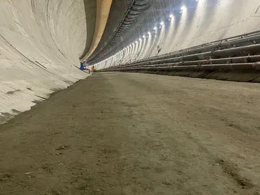 Wnętrze budowanego tunelu o betonowych ścianach, oświetlonego szeregiem lamp, z rurami i instalacjami biegnącymi wzdłuż prawej ściany.