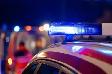 Nocna scena z bliskiego planu policyjnego lub strażackiego pojazdu z niebieskim światłem na dachu, w tle rozmyte światła służb ratunkowych.