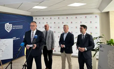 Czterech mężczyzn stoi podczas konferencji prasowej dotyczącej zintegrowanej sieci kolejowej, jeden z nich przemawia do mikrofonów.