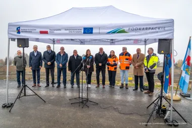 Grupa osób stoi pod namiotem promocyjnym z logotypami Podkarpacia i UE podczas plenerowej konferencji prasowej na terenie inwestycji.