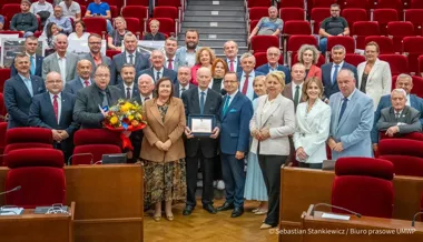 Grupa osób pozuje do pamiątkowego zdjęcia w auli z czerwonymi fotelami, jedna osoba trzyma bukiet kwiatów, a inna dyplom.