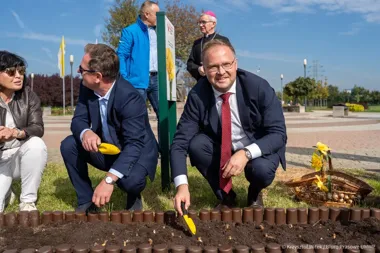 Kilka osób w eleganckim ubraniu sadzi cebulki kwiatowe w przygotowanej ziemi podczas uroczystości na otwartej przestrzeni miejskiej.