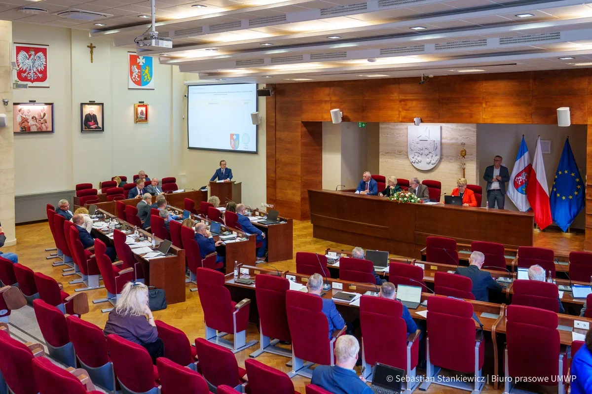 Posiedzenie w auli z czerwonymi fotelami, podczas którego mówca przemawia z pulpitu do uczestników siedzących przy biurkach z laptopami.